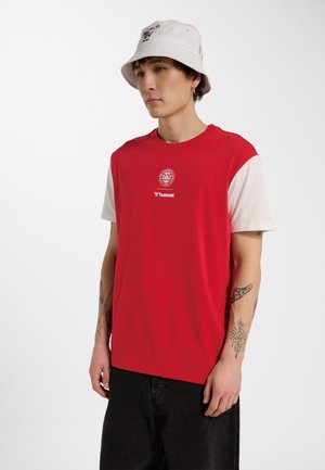 Mężczyzna ubrany w czerwoną i białą koszulkę z krótkim rękawem z logo na piersi, czarne spodnie, biały kapelusz typu bucket hat oraz widoczne tatuaże na ramionach.