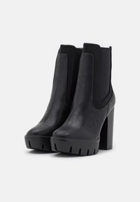 Bottes chevilles noires avec une tige élégante en cuir synthétique, des panneaux latéraux élastiques et un talon épais. La semelle présente une bande de roulement texturée pour une meilleure adhérence.