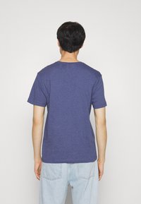 Mand stående med ryggen til iført en ensfarvet blå kortærmet T-shirt og lyseblå jeans mod en hvid baggrund.