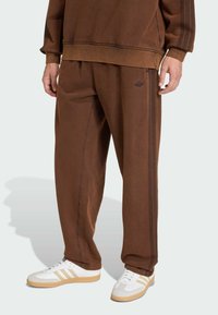 Person in braunen Adidas Jogginghosen und passendem Sweatshirt mit weißen Sneakers vor einfarbigem Hintergrund.