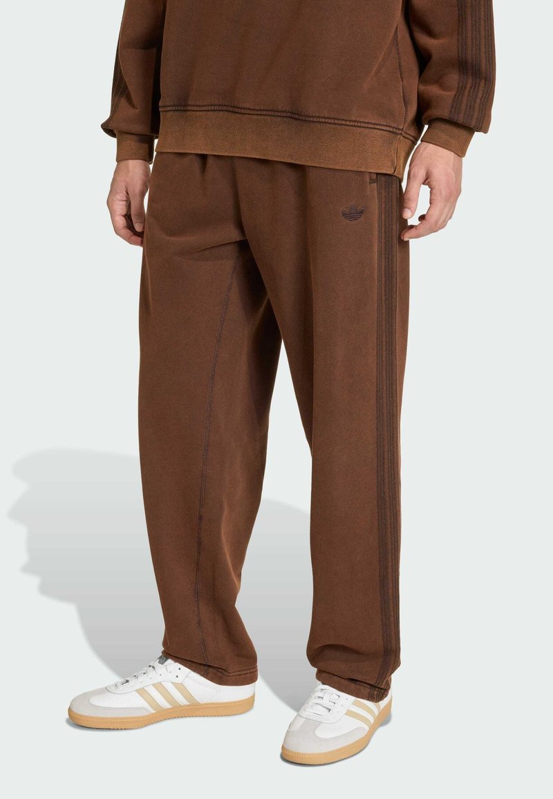 Person in braunen Adidas Jogginghosen und passendem Sweatshirt mit weißen Sneakers vor einfarbigem Hintergrund.