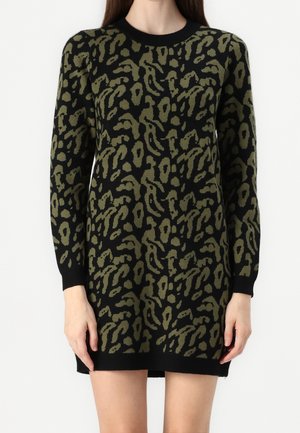 Długa czarna sukienka z zielonym oliwkowym abstrakcyjnym wzorem w cętki, dopasowanymi mankietami i krawędzią kończącą się na środku uda, noszona przez stojącą osobę.