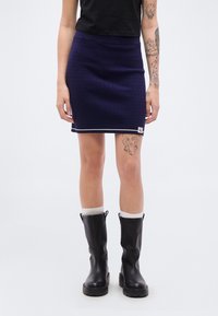 Πλεκτή μίνι φούστα σε ναυτικό μπλε με υφασμένο σχέδιο, αντίθετο λευκό τελείωμα και πλαϊνή ετικέτα με λογότυπο, συνδυασμένη με μαύρες μπότες έως το γόνατο και λευκές κάλτσες.