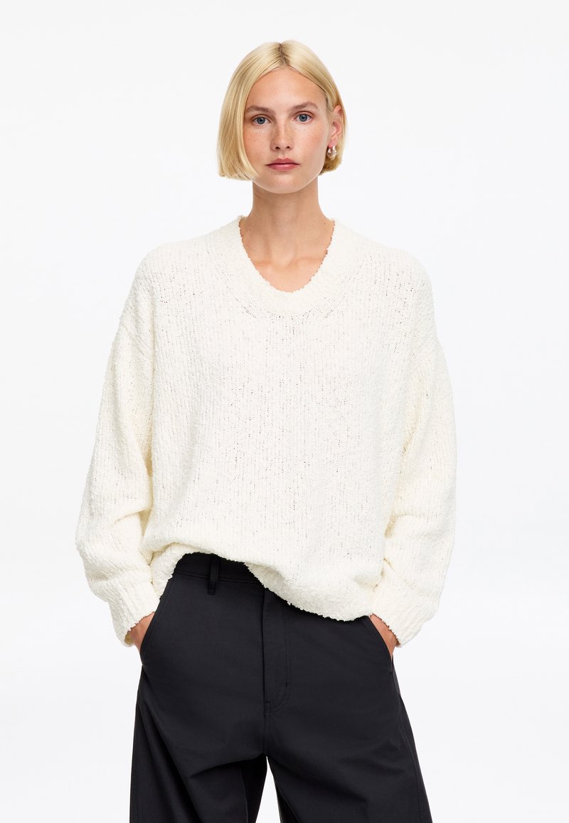 Femme aux cheveux blonds courts portant un pull en maille blanc ample et un pantalon noir, debout les mains dans les poches sur un fond blanc uni.