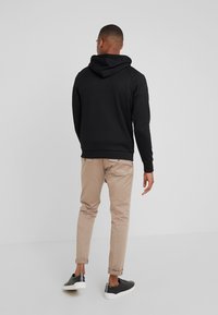 Man som bär en svart hoodie, beige uppvikta byxor och svarta sneakers, står med ryggen mot kameran på en enfärgad grå bakgrund.
