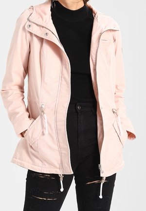 Vrouw draagt een lichtroze jack met ritssluiting over een zwart geribd shirt en zwarte gescheurde spijkerbroek, handen in de zakken van het jack.