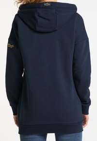 Mörkblå hoodie med en stor bakhuva, ribbade ärmslut och nederkant. Har en diskret logotyp i guld på vänster ärm. Tillverkad av mjukt bomullsmaterial.