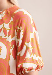 Satin blouse met een gedurfde roze en gele bloemenpatroon, met puffed mouwen en gegatherde manchetten, op een witte achtergrond.