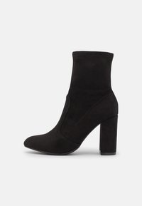 Bottine en daim noir avec bout pointu, talon bloc haut, tige ajustée, design sans coutures, et panneaux latéraux élastiques pour un enfilage facile.