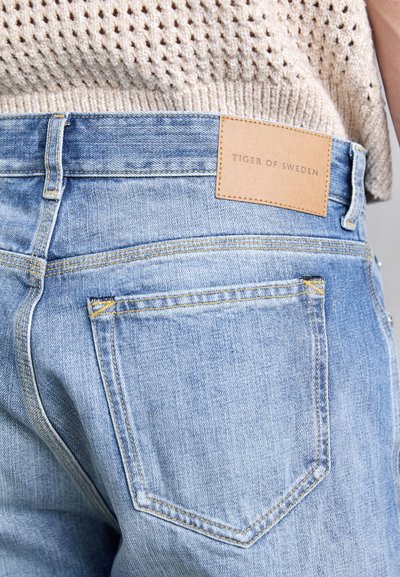 Jeans din denim de un albastru deschis au un etichetă din piele pe care este inscripționat "TIGER OF SWEDEN", cu buzunare la spate și accente de cusătură galben.