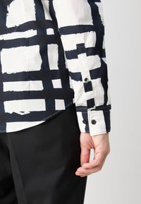 Chemise boutonnée présentant un motif abstrait de coups de pinceau bleu marine et blanc, confectionnée dans un tissu léger avec des poignets boutonnés et un ourlet arrondi.
