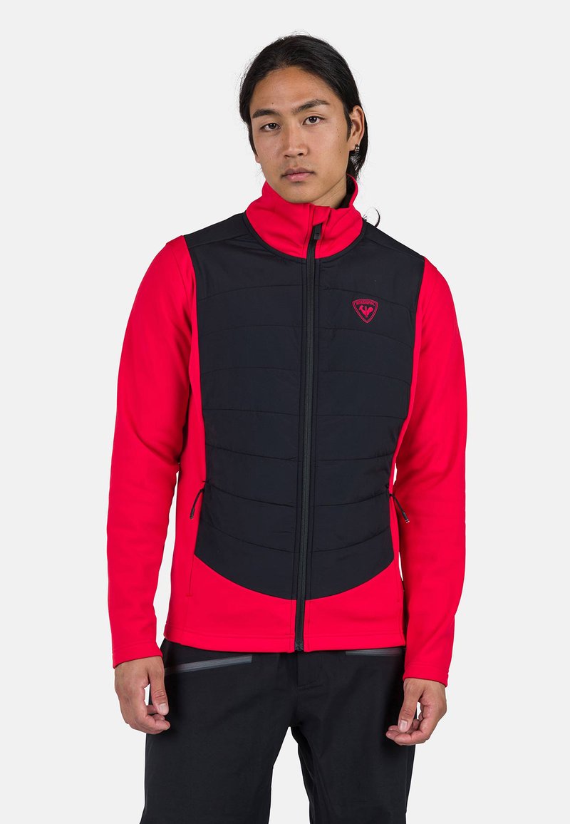 Červená a černá zateplená vesta se zipem vpředu, polstrovanou oblastí hrudi a bočními kapsami. Vesta má vysoký límec a logo na hrudi.