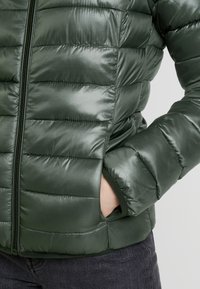 Polstret grøn jakke med en blank finish, der har vandret quiltning, en frontlynlås og en synlig sidelomme.