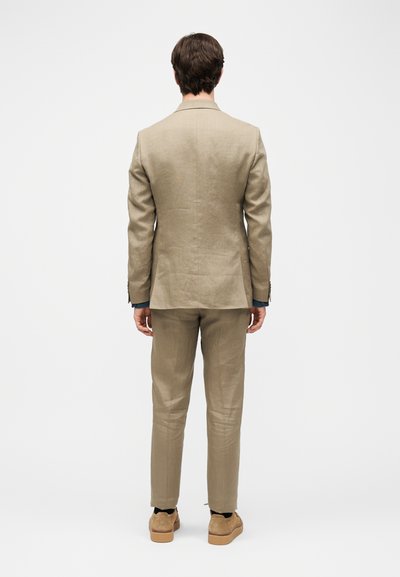 Άνδρας που στέκεται με την πλάτη, φοράει μπεζ κοστούμι με παντελόνι ταιριαστό και ανοιχτό καφέ παπούτσια σε απλό λευκό φόντο.