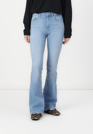 Donna che indossa jeans a vita alta svasati azzurri, maglia a maniche lunghe nera e scarpe con punta marroni con stampa leopardata, in piedi su pavimento bianco.