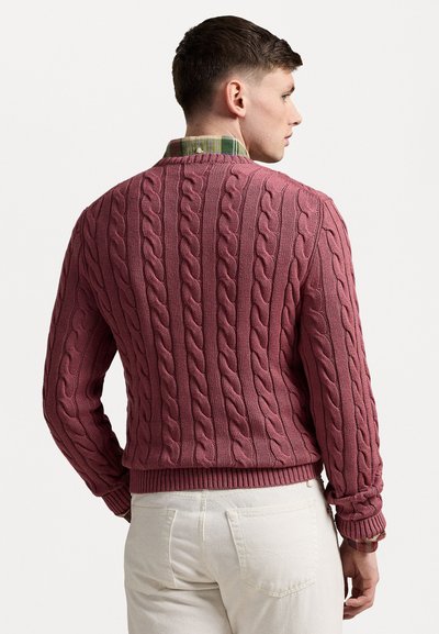 Cable-knit sweater in dusty rose. Features ribbed cuffs and hem, textured patterns, and layered over a green and plaid shirt.