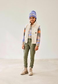 Fleece vest over een gestreepte trui, groene cargo broek en beige laarzen. Blauwe gebreide beanie met letter patch. Zachte texturen, neutrale kleuren.