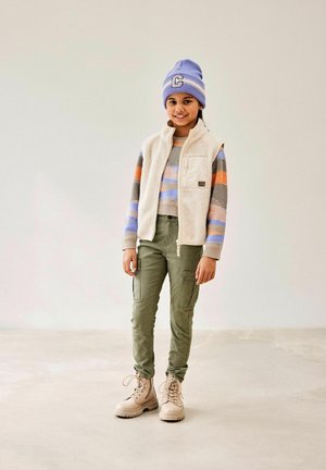 Gilet en polaire sur un pull rayé, pantalon cargo vert et bottes beiges. Bonnet en tricot bleu avec une écusson. Textures douces, couleurs neutres.