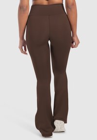 Donna in piedi che indossa pantaloni a zampa di elefante a vita alta di colore marrone scuro e scarpe da ginnastica bianche, vista da dietro su uno sfondo semplice.