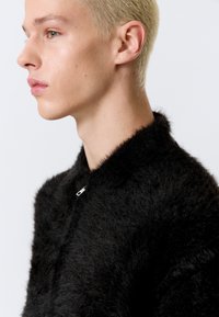 Μαύρο, αφράτο μπουφάν με στρογγυλό γιακά και μικρή ασημένια λεπτομέρεια. Κοντινή λήψη που αναδεικνύει την υφή και το σχέδιο του γιακά.