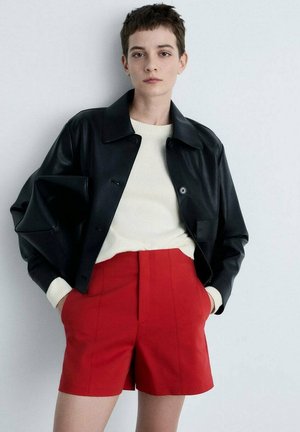 Black leather jacket over a cream sweater and red high-waisted shorts; features a collared design, buttons, and ribbed cuffs.