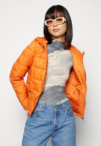 Casaco puffer laranja com capuz e design matelassado, usado sobre uma blusa cinza estampada. Combinado com jeans de cintura alta azul claro.