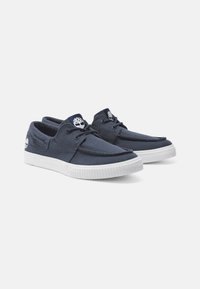 Marineblaue Canvas-Sneaker mit einer weißen Gummisohle, die ein klassisches Bootsschuh-Design aufweisen, mit gestickten Details und einem Logo auf der Zunge und der Seite.