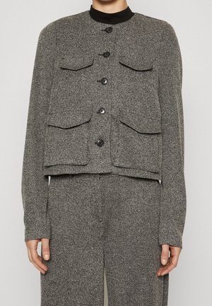 Femme portant une veste grise texturée boutonnée avec quatre poches à rabat à l'avant, sur un pantalon large assorti.