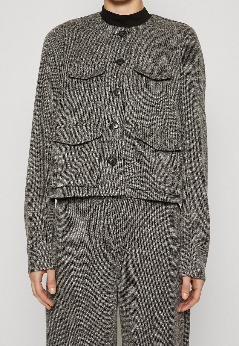 Femme portant une veste grise texturée boutonnée avec quatre poches à rabat à l'avant, sur un pantalon large assorti.