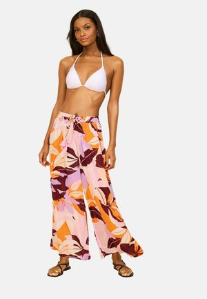 Donna che indossa un top bikini bianco e pantaloni a gamba larga con stampa di foglie tropicali arancioni, rosa e marroni, in piedi su uno sfondo semplice.