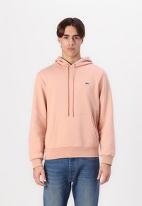Sudadera rosa claro con bolsillo tipo canguro, cordones y un pequeño logo verde de Lacoste en el pecho. Tejido suave, con puños y dobladillo acanalados.