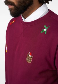 Bordeaux sweatshirt med broderade detaljer: en katt i tomteluva, en kvist av mistel och dekorativa godiskäppar. Mjuk bomullstextur.