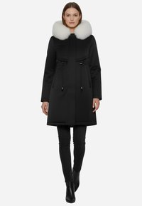 Parka nera con colletto in pelliccia bianca staccabile, dotata di lacci in vita, chiusura con zip e tasche frontali. Tessuto liscio.