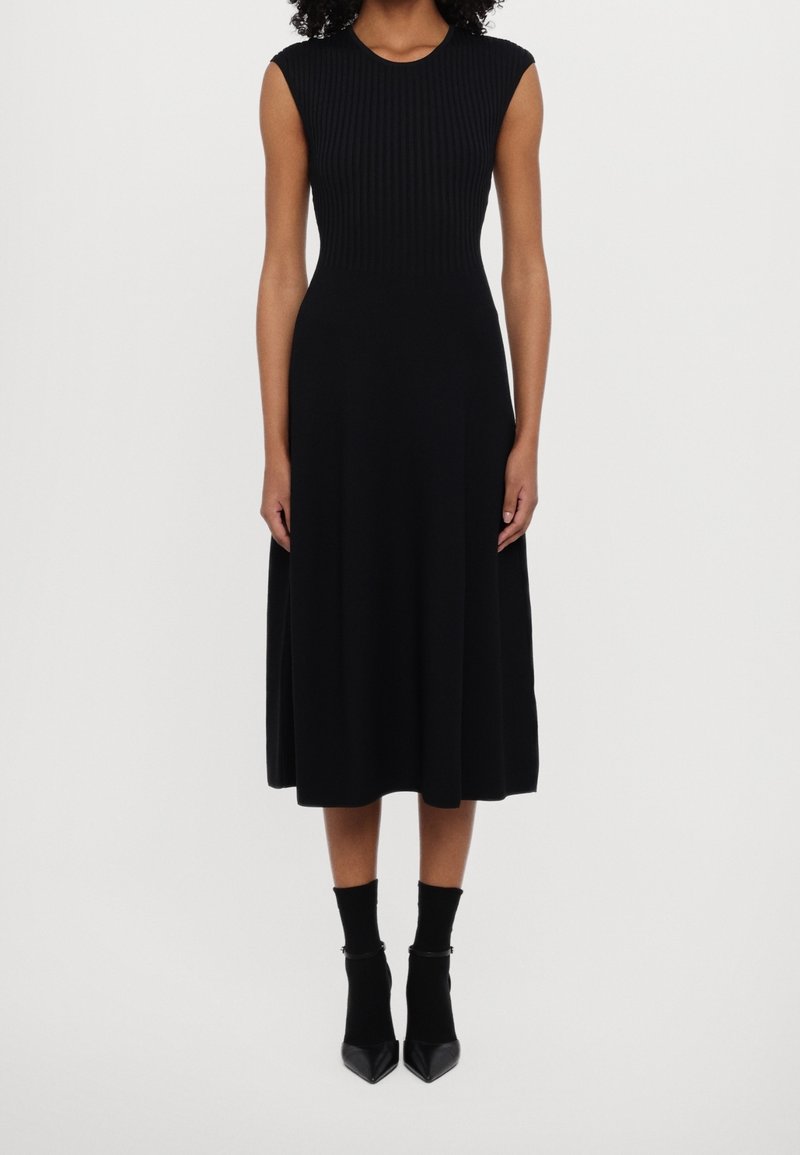Femme portant une robe midi noire sans manches avec un haut côtelé et une jupe évasée, associée à des bottines noires et des chaussettes.