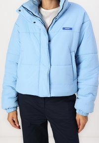 Světle modrá péřová bunda s krátkým střihem, s elastickými manžetami a předním zapínáním na cvoky. Logo nášivka u límce.