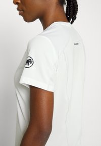 Perfil de una persona que lleva una camisa de manga corta blanca con un logo de mamut negro en la manga, contra un fondo liso.