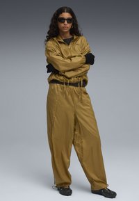 Tuta leggero verde oliva con giacca con zip e pantaloni con vita elastica. Texture opaca, presenta un logo e guanti e scarpe nere.