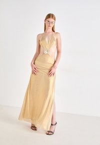 Goldsatin-Kleid mit tiefem Ausschnitt, Ausschnitten an der Taille und einem Blumenakzent. Das Model trägt schwarze Absatzsandalen und eine Brille.