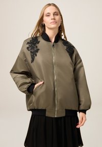 Giacca bomber verde oliva con accenti neri ricamati sulle spalle, chiusura con zip, polsini e orlo a costine, tasche laterali e finitura satinata.