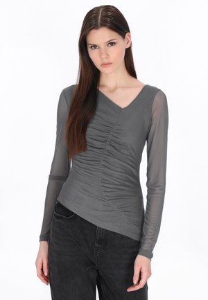 Młoda kobieta z długimi brązowymi włosami w dopasowanym szarym marszczonym topie z przezroczystymi długimi rękawami i czarnych spodniach, stojąca na białym tle.