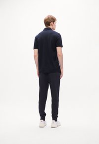Joven de pie de espaldas, vistiendo una camiseta polo negra, pantalones jogger negros y zapatillas blancas sobre un fondo blanco.