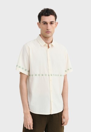 Junger Mann, der ein cremefarbenes Kurzarmhemd mit grünen Sternmustern auf Brust und Ärmeln trägt und vor einem einfarbigen Hintergrund steht.