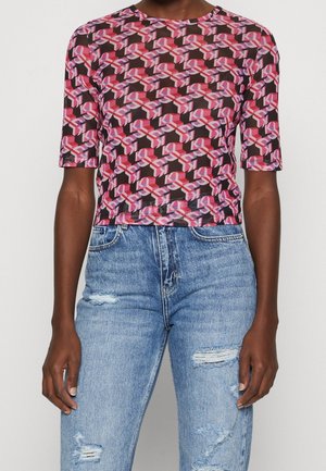 Personne portant un haut à manches courtes à motifs géométriques rose, rouge et violet avec un jean bleu déchiré à taille haute.