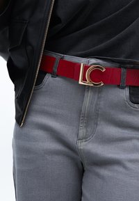 Červený lesklý opasek se zlatou sponou 'LC', nošený s šedými džíny a černou bundou. Důraz na elegantní design a kontrastní barvy.