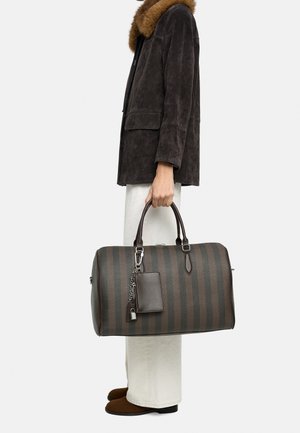 Osoba w kurtce z ciemnobrązowym futrzanym kołnierzem i białych spodniach trzyma dużą podróżną torbę w ciemnobrązowe paski z przyczepioną skórzaną metką.