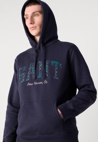 Felpa blu navy con tasca a marsupio, caratterizzata da ricamo "GANT" in plaid trapuntato e la scritta "New Haven, Ct." sul petto.