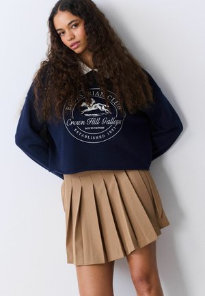 Kvinde med langt krøllet hår iført en marineblå sweatshirt med ride-logo og et beige plissert skørt, stående op ad en hvid væg.