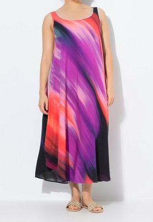 Femme portant une robe fluide sans manches avec un motif abstrait vibrant rouge, violet et noir, associée à des sandales beige ornées de strass.