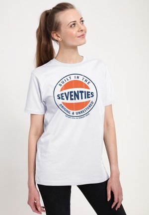 Ung kvinde med hestehale iført en hvid t-shirt med teksten "Built in the Seventies, Original & Unrestored" i orange og blå.