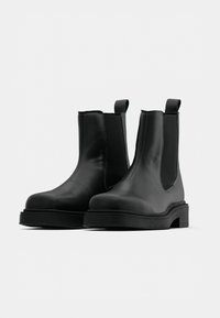 Mustat nahkaiset Chelsea-nilkkurit, joissa on joustavat sivupaneelit, pyöristetty kärki, vetonauhat ja paksut pohjat. Sileä pinta, jossa näkyviä ompeluyksityiskohtia.